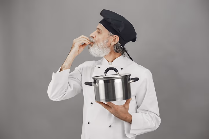Chef en uniforme goûtant un plat avec une marmite de cuisson, exprimant sa satisfaction par un geste de la main.